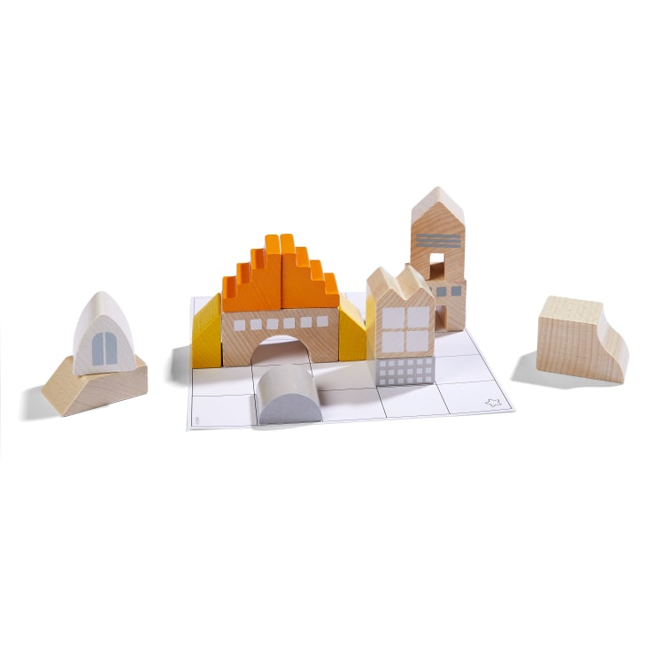 Mehrere Holzbausteine in verschiedenen Formen und Farben, darunter Gelb, Orange, Grau und Naturholz, sind auf einer weißen Gittermatte so angeordnet, dass sie Gebäuden und Bögen ähneln.