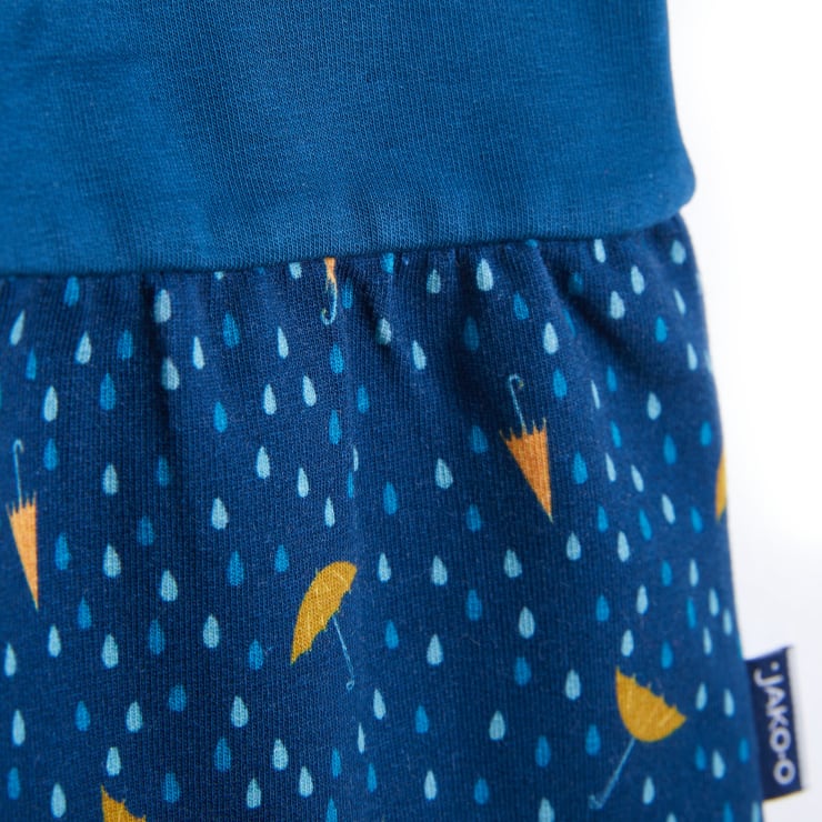 Nahaufnahme eines dunkelblauen Stoffes mit einem Muster aus kleinen blauen Regentropfen und gelben Regenschirmen, mit einer sichtbaren Naht und einem blauen Etikett auf der rechten Seite.