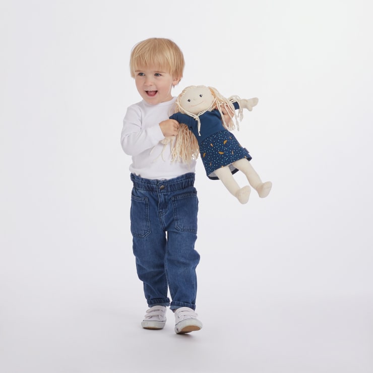 Ein kleines Kind mit blondem Haar, das ein weißes langärmeliges Hemd, blaue Jeans und weiße Turnschuhe trägt, hält eine Stoffpuppe in einem blauen Outfit, während es vor einem schlichten weißen Hintergrund steht.
