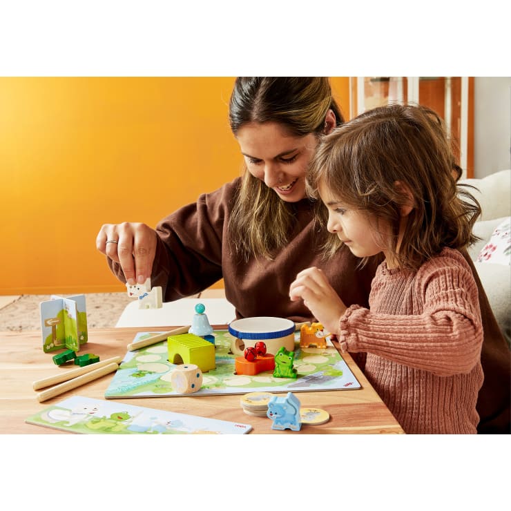 Ein Erwachsener und ein Kind sitzen an einem Tisch und spielen ein Brettspiel mit Tierfiguren und bunten Spielsteinen. Im Hintergrund sind eine orangefarbene Wand und eine beigefarbene Couch zu sehen.