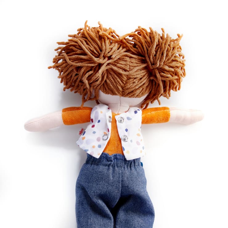 Auf einem weißen Hintergrund liegt eine Stoffpuppe mit zu zwei Puscheln gestyltem Garnhaar, die eine weiße Weste mit bunten Punkten, ein orangefarbenes Hemd und eine blaue Hose trägt.