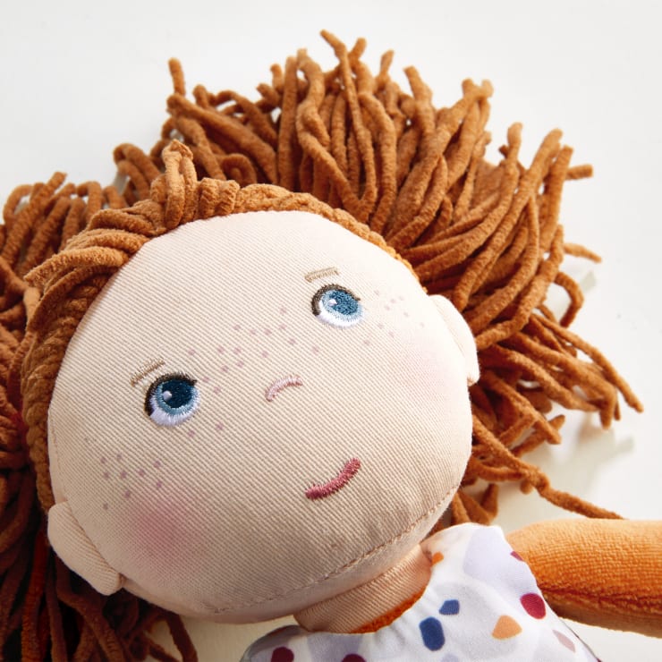 Nahaufnahme einer Stoffpuppe mit braunem Garnhaar, blauen gestickten Augen, ein paar Sommersprossen und einem gemusterten Kleid, die auf einer weißen Fläche liegt.