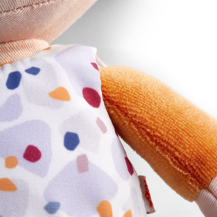 Nahaufnahme eines Plüschtiers, das einen weißen Stoffanzug mit einem bunten geometrischen Muster trägt und einen Teil seines orangefarbenen Arms und seiner Seite vor einem weißen Hintergrund zeigt.