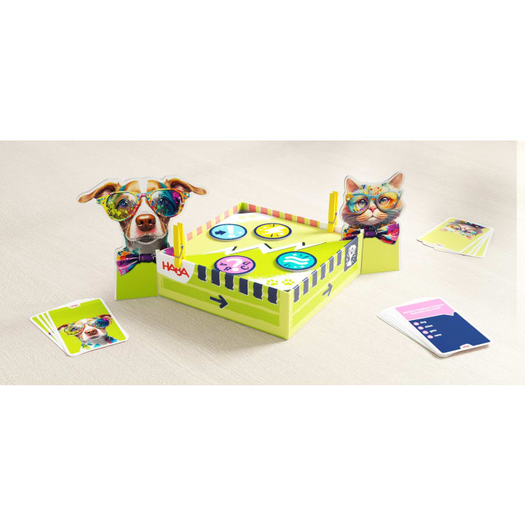 Ein farbenfrohes Tischspiel mit Pappfiguren eines Hundes und einer Katze mit Brille und Fliege, einem kleinen quadratischen Spielbrett mit Symbolen und mehreren Kartendecks auf einer hellen Oberfläche.