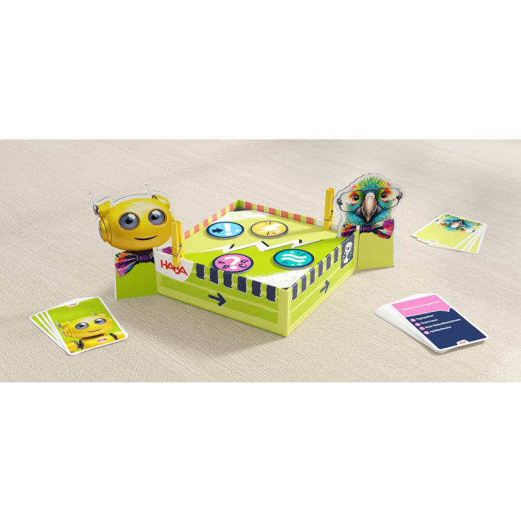Ein farbenfrohes Tischspiel mit einer quadratischen Papparena mit Cartoon-Roboter- und Papageienfiguren an zwei Ecken, Spielkarten und drei runden Plättchen mit verschiedenen Symbolen in der Mitte.
