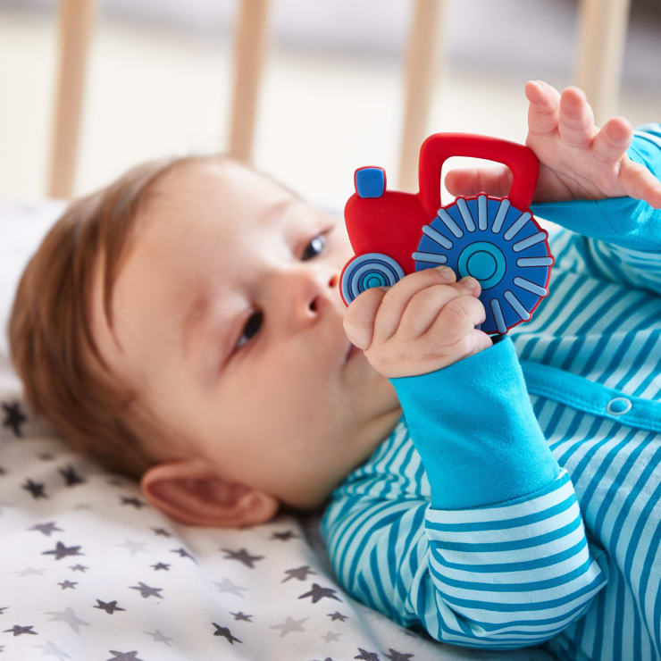 Ein Baby, das in einem blau-weiß gestreiften Strampler auf einem Sternchentuch liegt, hält einen rot-blauen Spielzeugtraktor und inspiziert ihn.