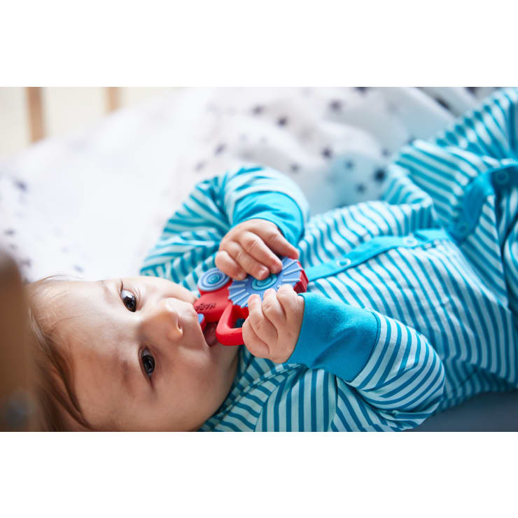 Ein Baby in einem blau gestreiften Strampler liegt auf einem Bett und kaut auf einem rot-blauen Spielzeug, während es nach oben schaut. Im Hintergrund ist eine weiße Decke mit blauen Mustern zu sehen.