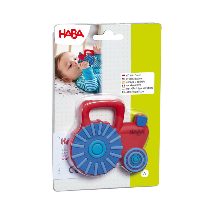 Ein rot-blaues Spielzeug in Form eines Traktors ist auf einer Pappunterlage verpackt. Auf der Verpackung sind das HABA-Logo und das Foto eines Babys zu sehen, das das Spielzeug in den Mund nimmt. Der Text ist in mehreren Sprachen aufgedruckt.