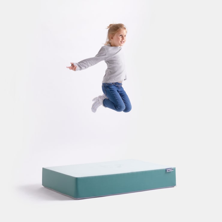 Ein kleines Kind in einem grauen Pullover und blauen Jeans springt in der Luft auf einer hellblauen rechteckigen Schaumstoffmatte vor einem schlichten weißen Hintergrund.