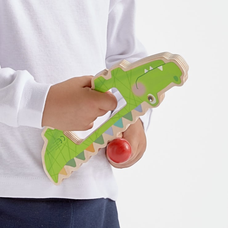 Ein Kind in einem weißen langärmeligen Hemd hält ein grünes Holzkrokodil mit einem roten Ball in der Hand. Der Hintergrund ist einfarbig weiß.