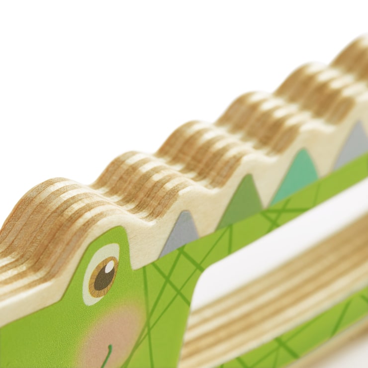 Nahaufnahme eines Holzspielzeugs, bemalt mit einem grünen, lächelnden Krokodilgesicht und einem zickzackförmigen, strukturierten Rand, der dem Rücken des Krokodils ähnelt. Das Spielzeug hat eine geschichtete Holzkonstruktion und geometrische Muster.