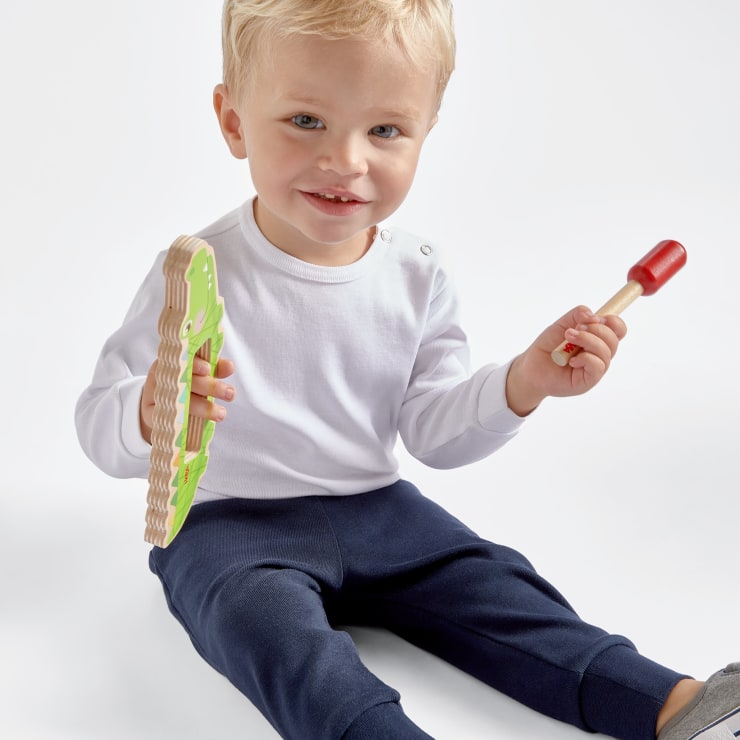 Ein kleines Kind mit blondem Haar sitzt auf dem Boden und hält ein grünes Holzspielzeug in der einen und einen roten Holzhammer in der anderen Hand. Es trägt ein weißes, langärmeliges Hemd und eine marineblaue Hose vor einem schlichten Hintergrund.