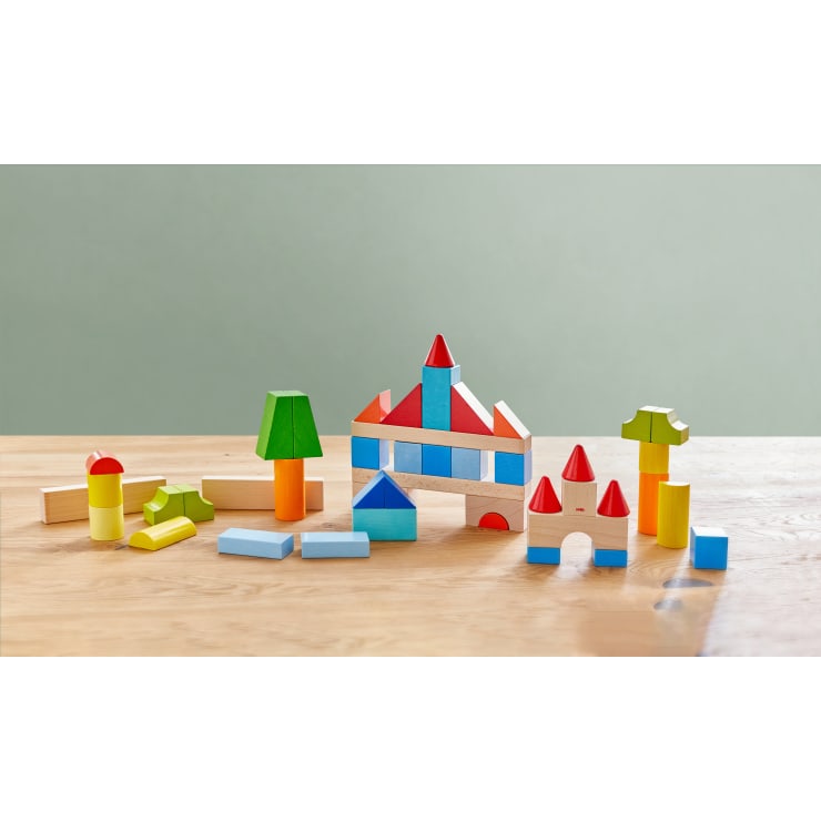 Bunte Holzbausteine sind auf einem hellen Holztisch angeordnet. Die Blöcke bilden verschiedene Strukturen, darunter Häuser, Bäume und einfache Formen, vor einem schlichten, hellgrünen Hintergrund.