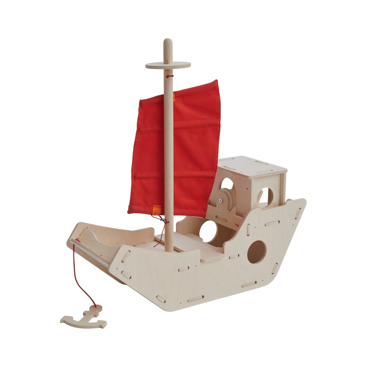 Ein hölzernes Spielzeugboot mit einem roten Stoffsegel, offenem Deck und einer kleinen geschlossenen Kajüte. Das Boot hat abgerundete Kanten und ausgeschnittene kreisförmige Fenster, an denen eine Zugschnur mit einem kleinen Holzanker befestigt ist.