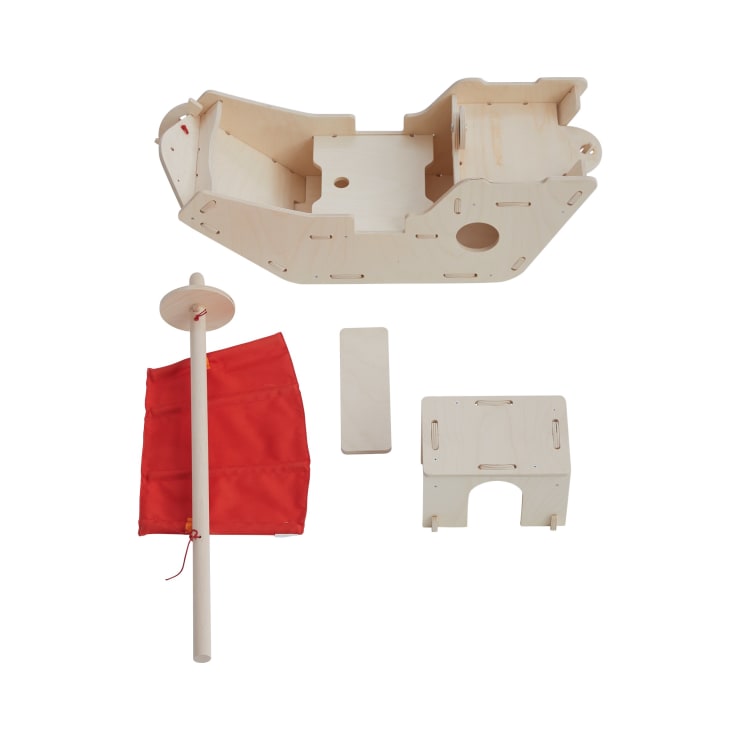 Ein zerlegter Bausatz für ein Holzspielzeugboot mit einem Hauptteil, einem rechteckigen Teil, einem kleineren Teil mit Ausschnitten und einem roten Stoffsegel, das an einem Holzdübel befestigt ist, alles auf einem weißen Hintergrund.