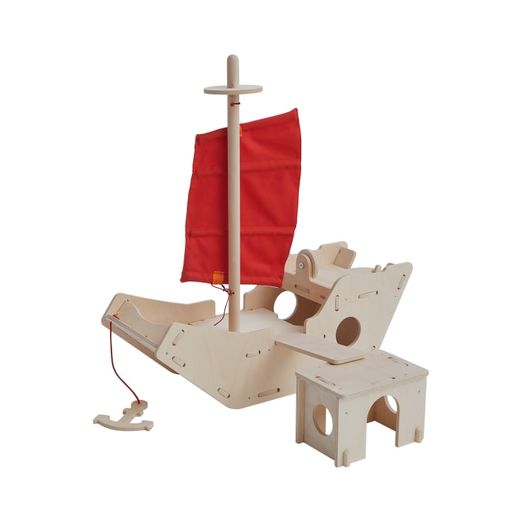 Ein Holzspielgerät in Form eines Bootes mit einem roten Stoffsegel, einem Steuerrad und einer kleinen Bank. Die Struktur ist aus leichtem Holz und hat ausgeschnittene Löcher und eine Zugschnur an der Vorderseite.