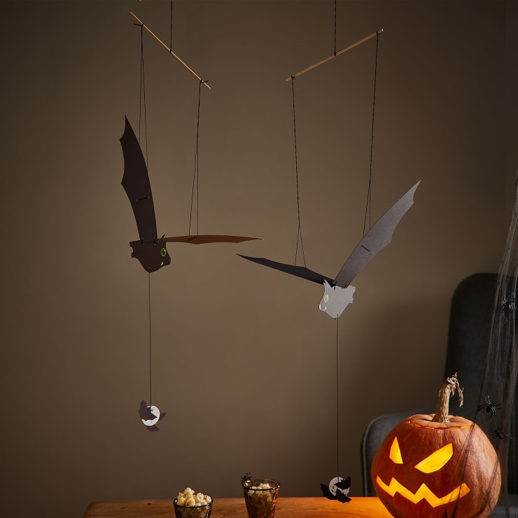 Zwei fledermausförmige Halloween-Mobiles hängen an schwarzen Schnüren an Stöcken. Jede Fledermaus hat einen Ausschnitt, der darunter hängt. Ein geschnitzter Kürbis mit einem leuchtenden Gesicht steht auf einem Tisch mit einer Schüssel Popcorn und einem Glas.
