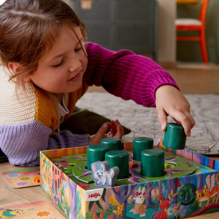 Ein kleines Kind liegt auf dem Boden, lächelt und spielt ein farbenfrohes Brettspiel mit großen grünen zylindrischen Spielsteinen und einer weißen Tierfigur auf dem Brett. Die Umgebung besteht aus einem Teppich und Möbeln.