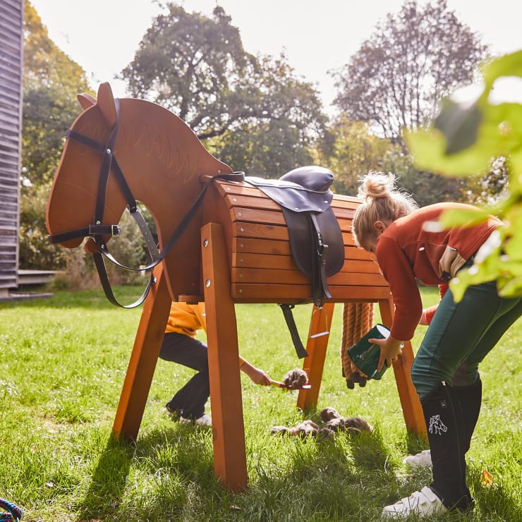 Zwei Personen räumen Gegenstände im Gras neben einem großen hölzernen Spielzeugpferd mit Sattel und Zaumzeug im Freien mit Bäumen und Sonnenlicht im Hintergrund auf.