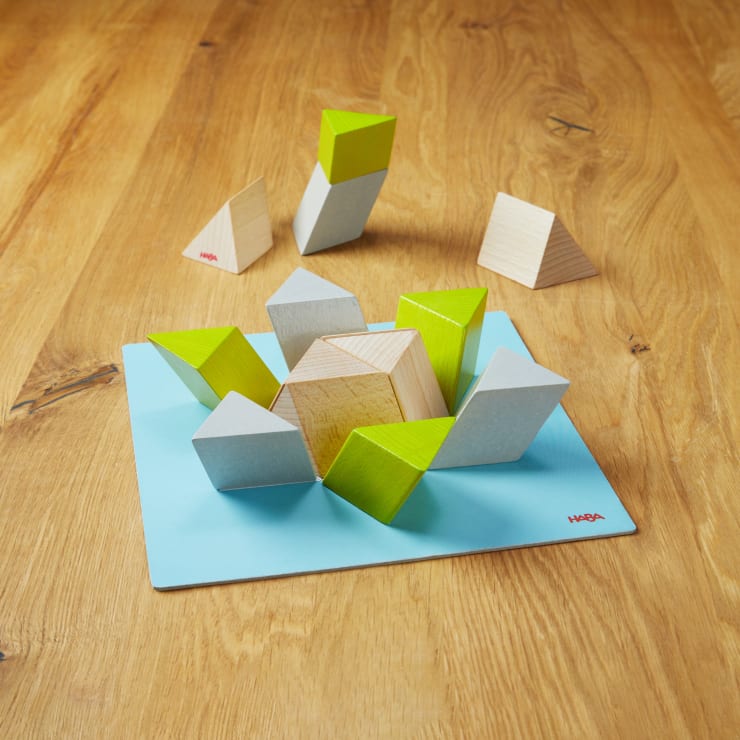 Mehrere geometrische Holzklötze, von denen einige grün und grau bemalt sind, sind auf einer blauen Matte auf einer Holzfläche angeordnet. Weitere Klötze sind im Hintergrund platziert. Die Matte hat in der Ecke das HABA-Logo.