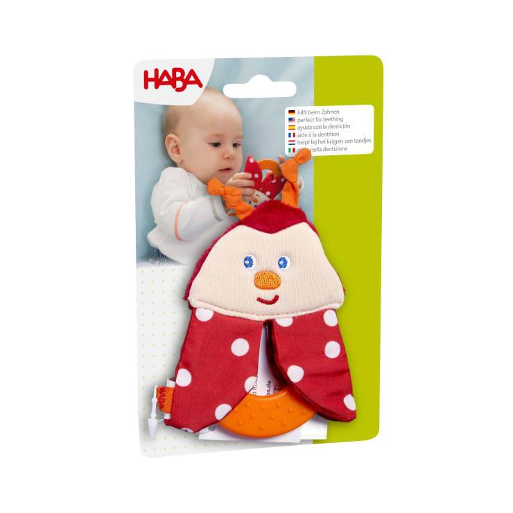 Ein verpacktes Babyspielzeug in Form eines rot-weiß gepunkteten Marienkäfers mit einem lächelnden Gesicht. Das Spielzeug ist auf einer Pappunterlage befestigt, die in der oberen linken Ecke ein Baby zeigt, das mit demselben Spielzeug spielt.