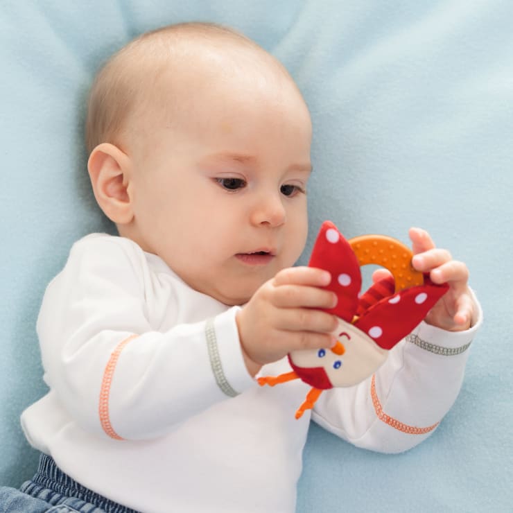 Ein Baby, das auf einer hellblauen Decke liegt, hält ein rot-orangefarbenes Spielzeug mit einem lächelnden Gesicht und gepunkteten Flügeln und untersucht es.