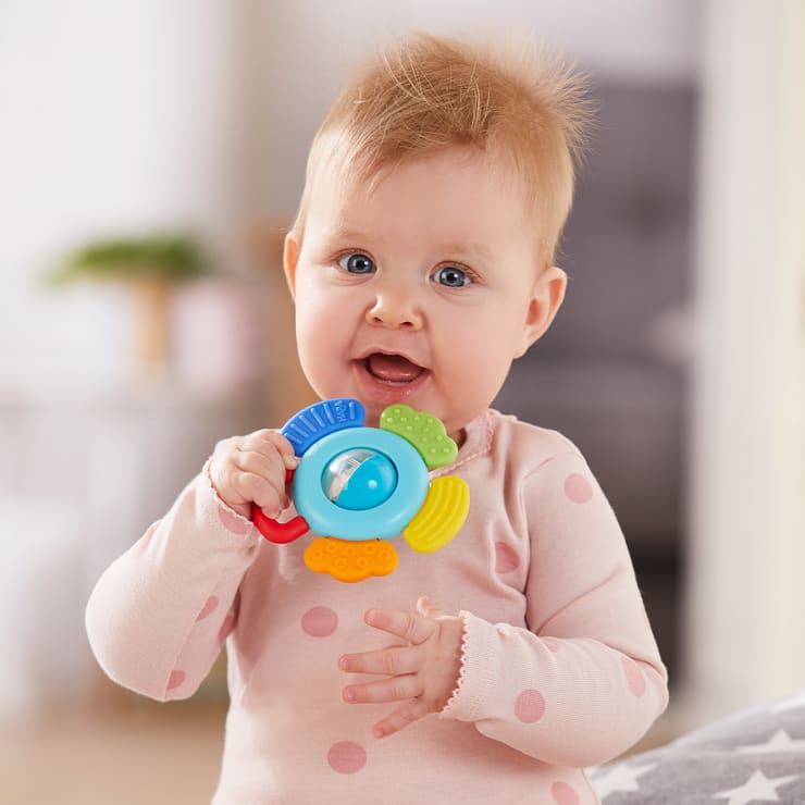 Ein Baby mit hellem Haar und einem rosa gepunkteten Strampler hält ein buntes Plastikspielzeug in Form eines Fisches und blickt mit offenem Mund in Richtung Kamera. Der Hintergrund ist sanft verschwommen.