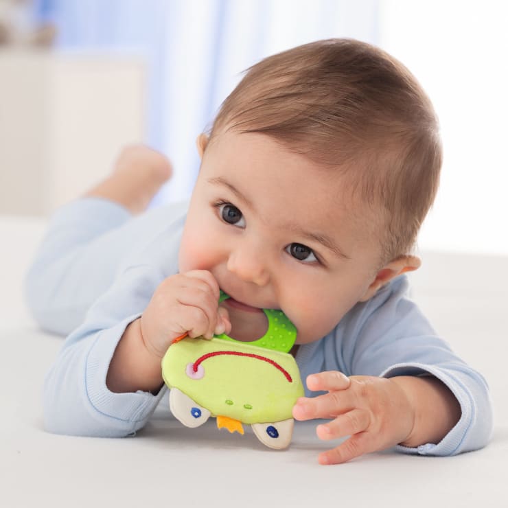Ein Baby im hellblauen Schlafanzug liegt auf dem Bauch und kaut auf einem grün-weißen Beißspielzeug herum. Der Hintergrund ist sanft in hellen Farben verschwommen.