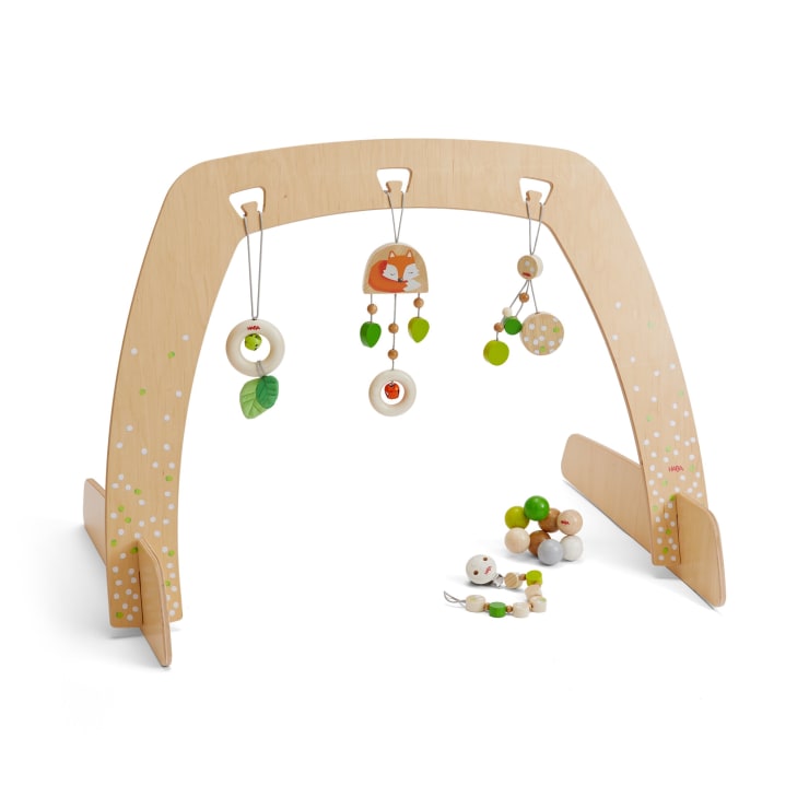 Ein hölzernes Babyspielgerät mit drei hängenden Spielzeugen in Grün-, Beige- und Orangetönen; eine Holzrassel und ein Beißring liegen auf dem Boden unter dem Bogen. Die Turnhalle hat kleine weiß und grün bemalte Details.