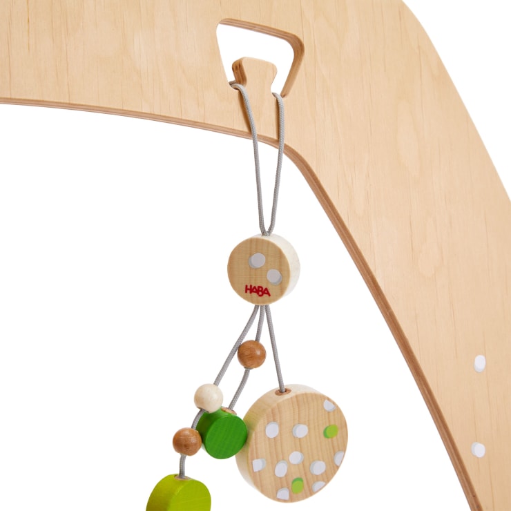 Nahaufnahme eines hölzernen Babytrainingsgeräts mit einem Hängespielzeug mit Holzperlen und -scheiben in natürlichen, grünen und weißen Farben, das an grauen Schnüren an einem Schlitz im Rahmen hängt.
