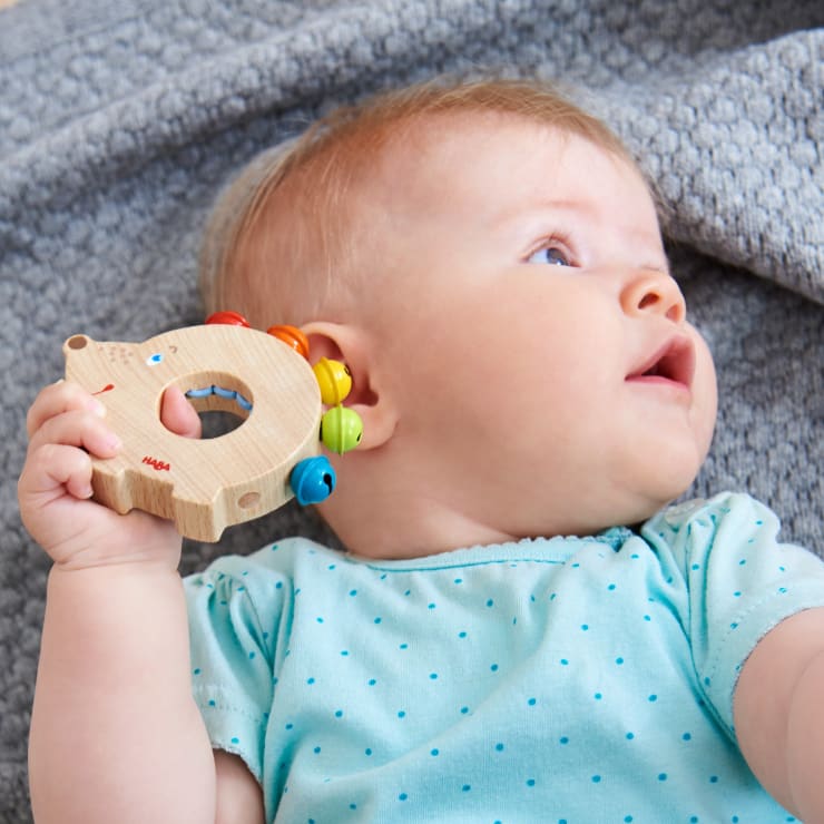 Ein Baby in einem hellblauen, gepunkteten Hemd liegt auf einer strukturierten grauen Decke, hält ein Holzspielzeug mit bunten Perlen in der Hand und schaut nach rechts.