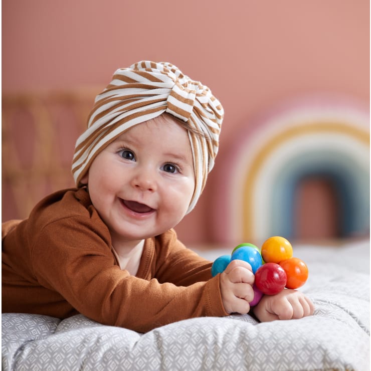 Ein Baby in einem braunen Outfit und einem gestreiften turbanartigen Stirnband liegt auf einem Bett, lächelt und hält ein buntes Rasselspielzeug. Im Hintergrund ist eine verschwommene Regenbogendekoration zu sehen.
