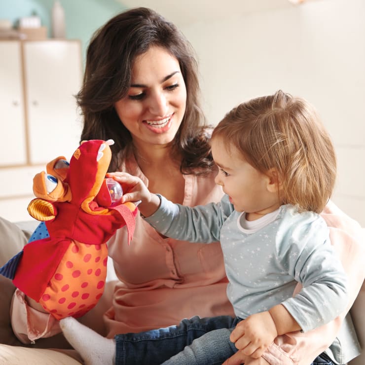 Eine Frau und ein kleines Kind sitzen zusammen in einem Haus. Die Frau hält eine bunte Handpuppe in der Hand, und das Kind streckt die Hand aus, um die Puppe zu berühren, während es lächelt. Sie scheinen beide beschäftigt und glücklich zu sein.