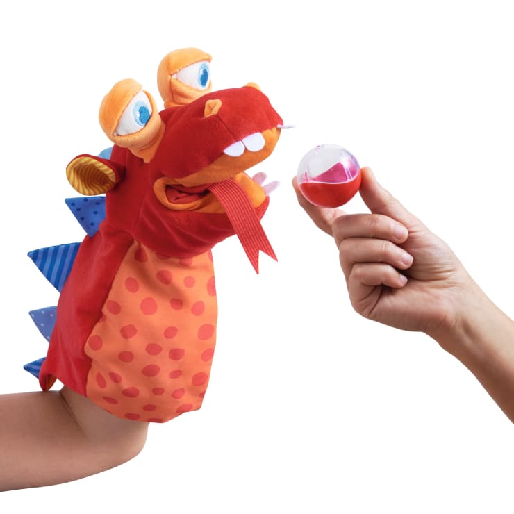 Eine Person hält eine rote und orangefarbene Drachenhandpuppe mit großen Augen und Filzzähnen, während eine andere Hand einen kleinen roten und durchsichtigen Plastikball vor die Puppe hält.