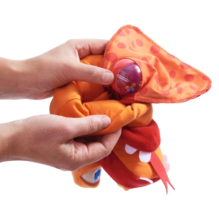 Zwei Hände halten und drücken ein orangefarbenes und rotes Plüschtier mit großen Schlappohren und einer durchsichtigen Rassel mit kleinen bunten Perlen im Inneren.