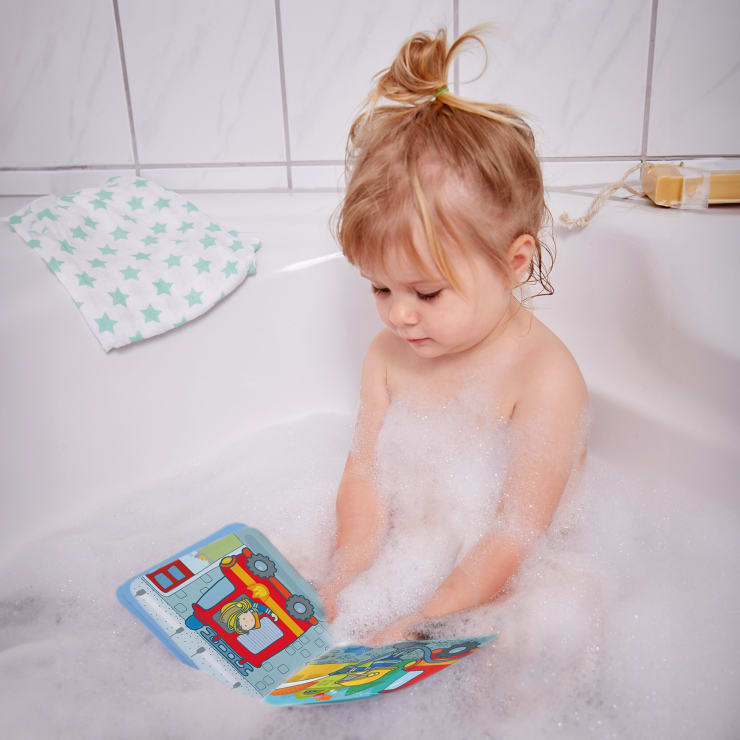 Ein kleines Kind sitzt in einer mit Seifenblasen gefüllten Badewanne und sieht sich ein buntes Buch an. Ein Waschlappen mit grünen Sternen liegt auf dem Wannenrand, und im Hintergrund sind weiße Kacheln zu sehen.