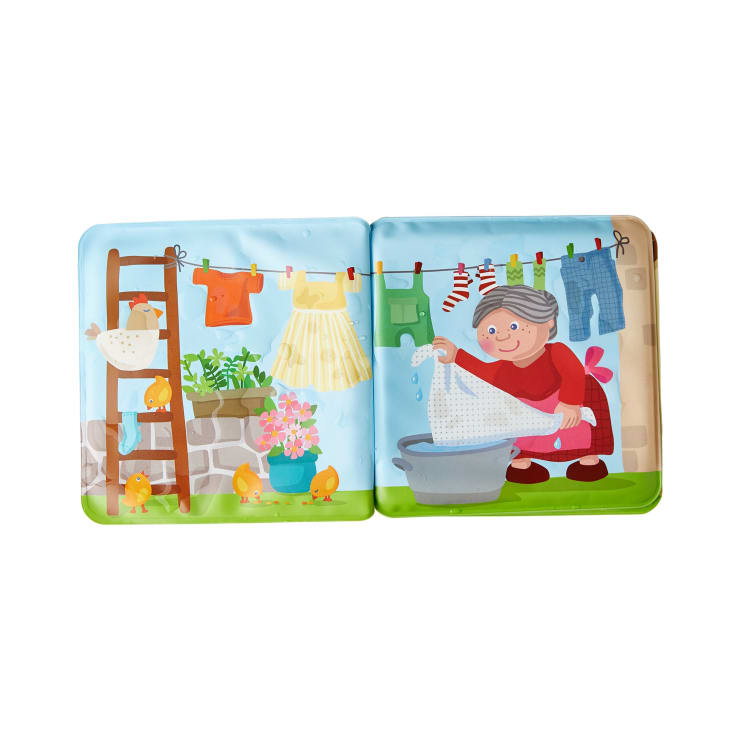 Eine illustrierte Kinderbuchseite zeigt eine ältere Frau, die im Freien in einer Wanne Wäsche wäscht. Die Wäsche hängt auf einer Leine, auf einer Leiter stehen ein Huhn und Küken, und im Hintergrund sind Pflanzen und Blumen zu sehen.