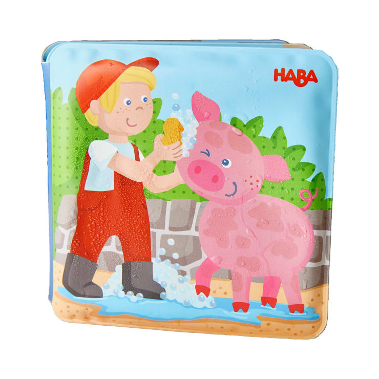 Ein Cartoon-Junge in roter Latzhose und Stiefeln wäscht ein rosa Schwein mit einem Schwamm. Wasser und Seifenblasen sind zu sehen. Der Hintergrund besteht aus Grünpflanzen und einer Steinmauer. Das Wort HABA ist oben rechts in Rot aufgedruckt.