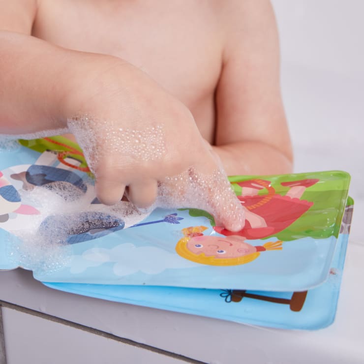 Die seifige Hand eines Kindes berührt ein wasserfestes Bilderbuch, während es in der Badewanne sitzt; das Buch zeigt bunte Cartoon-Illustrationen von Menschen und Szenen im Freien.
