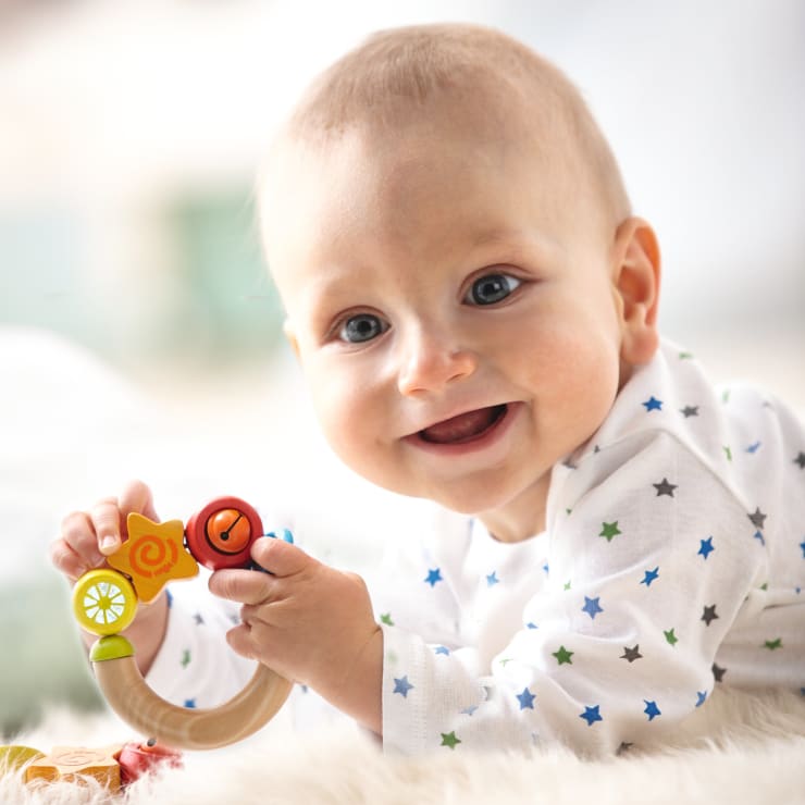 Ein Baby, das ein weißes Hemd mit blauen und grünen Sternen trägt, liegt auf einer weichen, weißen Oberfläche, lächelt und hält eine bunte Holzrassel in der Hand.