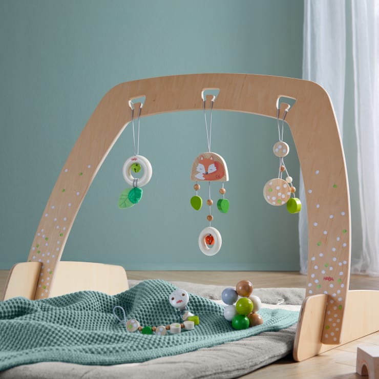 Ein hölzernes Babyspielgerät mit drei hängenden Spielzeugen steht über einer grünen, strukturierten Decke. Bunte Perlen und eine kleine Rassel liegen in der Nähe auf dem Boden. Das Zimmer hat eine hellgrüne Wand und einen durchsichtigen Vorhang.