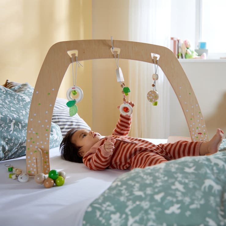 Ein Baby in einem rot-weiß gestreiften Strampler liegt auf einem Bett und greift nach Spielzeug, das an einem hölzernen Spielgerät hängt, umgeben von gemustertem Bettzeug in einem gut beleuchteten Raum.