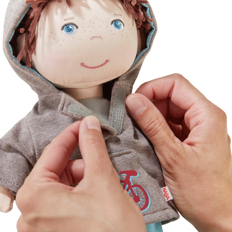Zwei Hände schließen den Klettverschluss an der grauen Jacke einer Stoffpuppe mit blauen Augen, hellbraunem Garnhaar und einer Fahrradgrafik auf der Jacke.