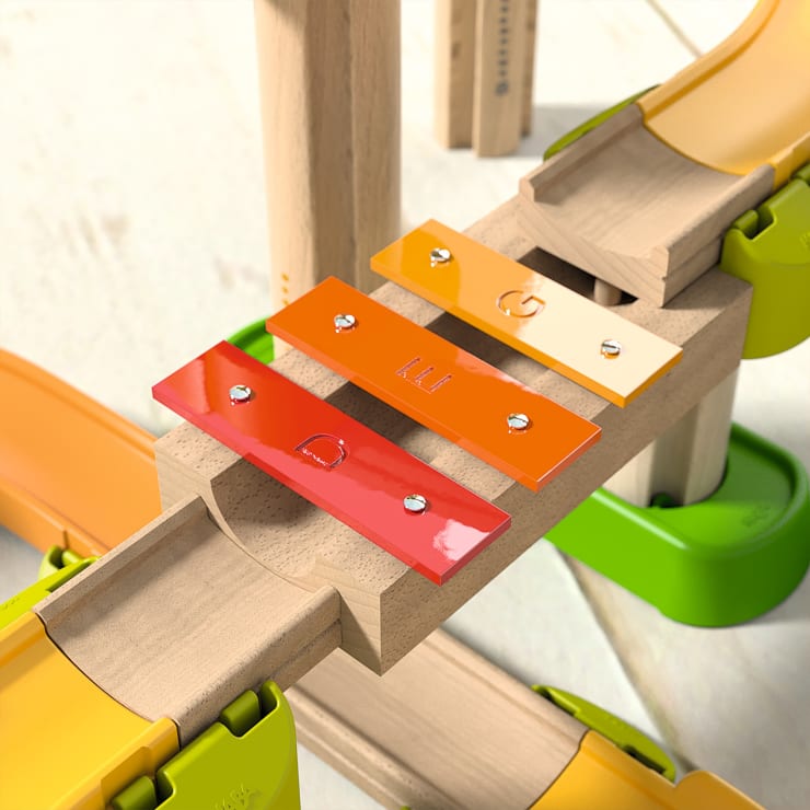 Nahaufnahme einer hölzernen Spielzeugeisenbahn mit drei xylophonartigen Metallstäben in Rot, Orange und Gelb, die auf einer Holzbrücke mit grünen und gelben Kunststoffstützen montiert sind.