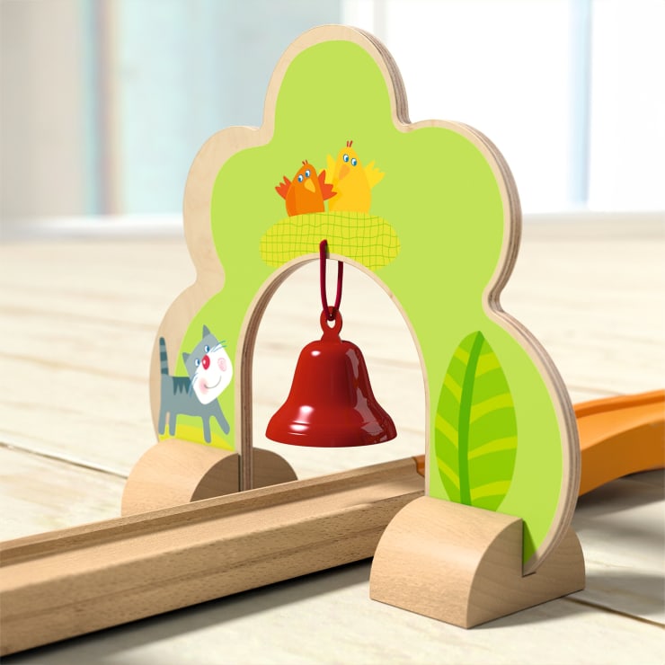 Ein hölzerner Spielzeugtorbogen mit einer roten Glocke in der Mitte. Der Bogen zeigt eine Cartoon-Katze, zwei Hühner in einem Nest und grüne Blätter, die auf einem hellen Holzboden mit Spielzeugeisenbahnschienen darunter liegen.