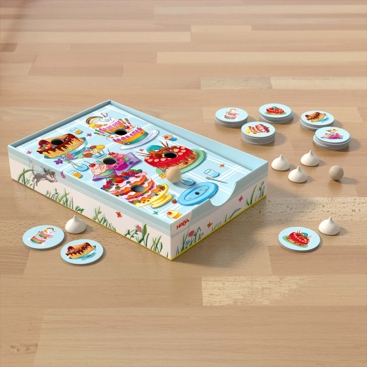Eine Brettspielschachtel für Kinder mit Cartoon-Desserts und Insekten auf dem Deckel steht auf einem Holzboden. Runde, illustrierte Spielsteine und weiße, kegelförmige Spielfiguren sind neben der Schachtel angeordnet.
