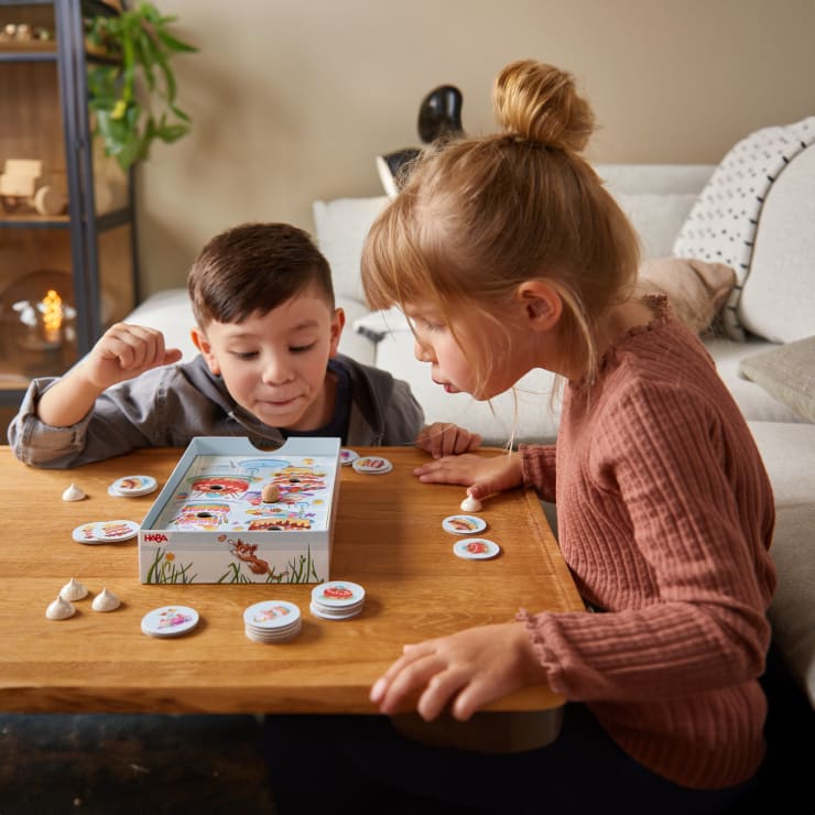 Zwei kleine Kinder sitzen an einem Holztisch und spielen konzentriert ein Brettspiel mit runden Figuren. Spielfiguren und Karten sind ausgebreitet, und im Hintergrund ist eine Couch mit Kissen zu sehen.