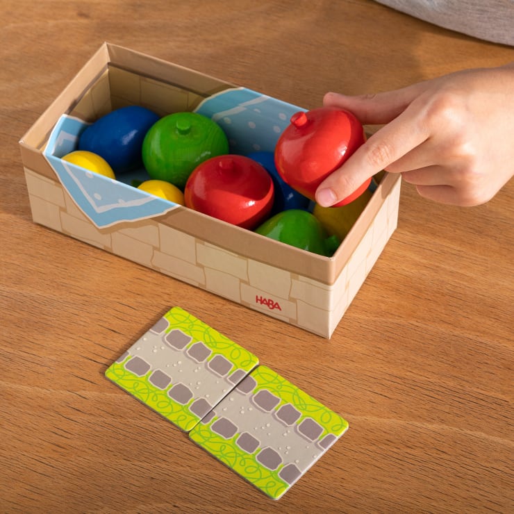 Eine Hand nimmt einen roten Holzapfel aus einer Kiste mit anderen bunten Holzfrüchten, während zwei grüne und graue Fliesen auf einem Holztisch daneben liegen.