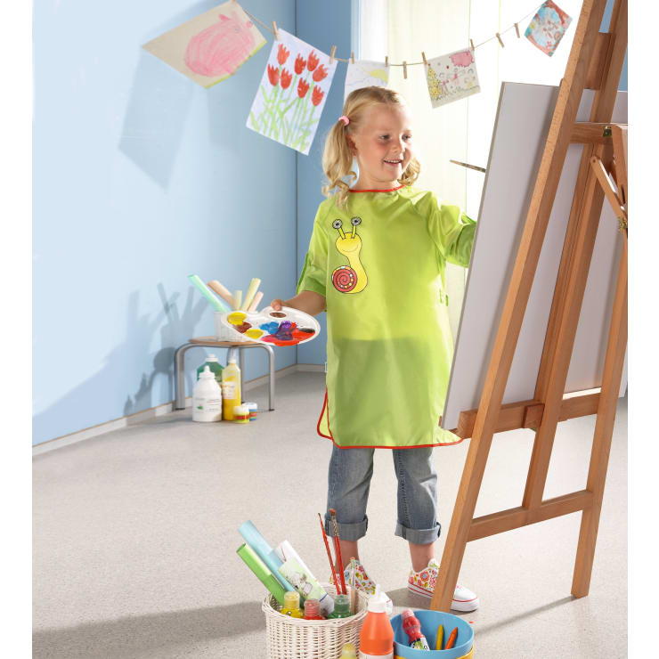 Ein junges Mädchen in einem grünen Kittel steht an einer Staffelei und malt auf einer Leinwand. In einem Korb auf dem Boden liegen Malutensilien, und im Hintergrund hängen Kinderzeichnungen an einer Wäscheleine.