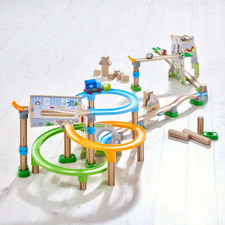 Ein hölzernes Murmelbahn-Spielzeugset mit bunten kreisförmigen Bahnen, Blöcken, Rampen, einem blauen Spielzeugauto, Murmeln und illustrierten Tafeln auf einem hellen Holzboden.